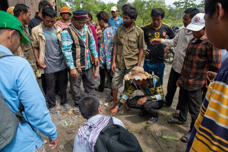 Upon arriving at the shrine within the concession, LASED III consultant King Vireaksith (foreground) began drawing boundaries and maps in the mud as Bunong community leader Lin Moa (traditional vest and cap) and other community members surrounded him to voice their concerns on July 14.(CamboJA/Andrew Califf)