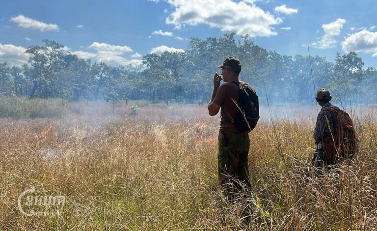 Ben Davis and a community ranger stand in a field of grass. A portion of the grass has been set on fire and smoke is visible. (CamboJA/ Sovann Sreypich)